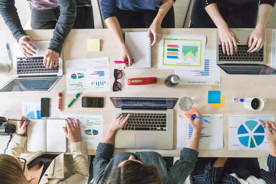 Overhead view of people working around a table with laptops, documents, charts, and stationery items.